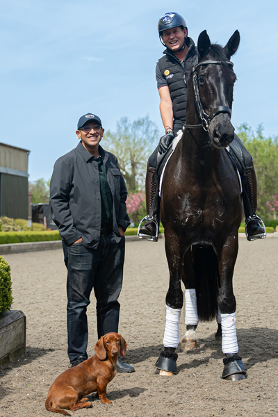 Equestrian rider on a black horse beside a person in dark clothing, with a small brown dog in front and greenery in the background